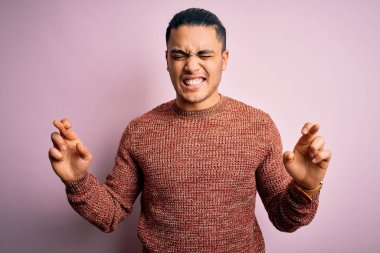 İzole edilmiş pembe arka planda dikilen sıradan süveter giyen genç Brezilyalı adam umut ve gözler kapalı gülümseyerek el sallıyor. Şans ve batıl inanç..