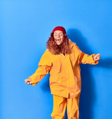 Genç, güzel, latin bir kadın balıkçı yağmurluğu giyiyor ve gülümsüyor. Yüzünde tebessümle zıplıyor, mavi arka planda yumruklarıyla kutlama yapıyor.