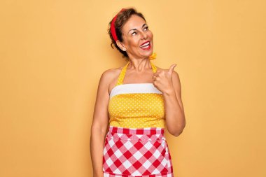 Orta yaşlı bir ev hanımı, 50 'li yılların modası tarzı retro elbise ve önlük giymiş mutlu yüzüyle gülümseyen ve baş parmağıyla yüzünü gösteren bir kadın..