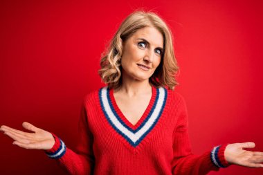 Orta yaşlı güzel sarışın kadın izole edilmiş kırmızı arka plan üzerine gündelik süveter giyiyor ve elleri ve kolları havada kafası karışmış bir ifade takınıyor. Şüphe kavramı.