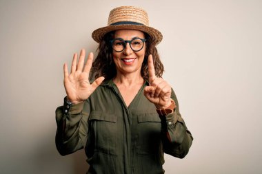 Orta yaşlı esmer kadın gözlüklü ve şapkalı. İzole edilmiş beyaz arka planda duruyor. Altıncı parmaklarıyla işaret ediyor..