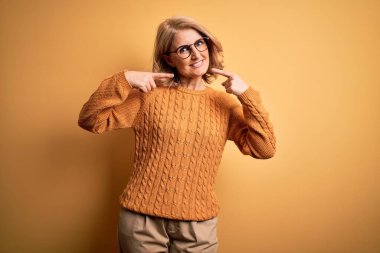 Sarışın, orta yaşlı, güzel bir kadın sarı arka planda gözlük takıyor. Gülümsüyor, parmaklarıyla ve ağzıyla işaret ediyor. Diş sağlığı konsepti.