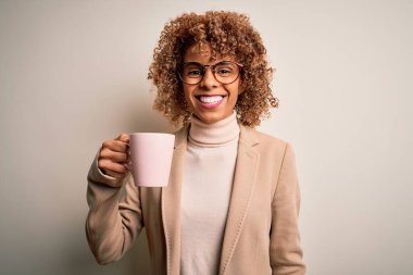 Genç Afro-Amerikan kıvırcık kadın izole edilmiş beyaz arka planda bir kupa kahve içiyor mutlu bir yüzle ayakta ve gülümsüyor dişlerini gösteren bir gülümsemeyle