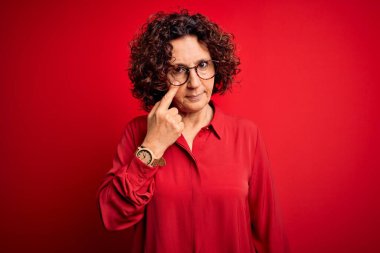 Orta yaşlı, kıvırcık saçlı, rahat gömlekli, kırmızı arka planda gözlüklü güzel bir kadın.