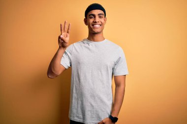 Yakışıklı Afro-Amerikan sporcu sarı arka planda spor giysisi giyiyor ve gülümserken üç numarayı işaret ediyor..