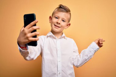 Küçük beyaz çocuk akıllı telefon kullanıyor ve telefonun ekranına bakıp mutlu bir şekilde elini ve parmağını yan tarafa gösteriyor.