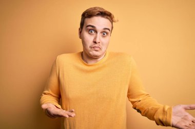 İzole edilmiş sarı arka plan üzerine sıradan süveter giyen genç, kızıl saçlı ve elleri ve kolları havada kafası karışmış bir adam. Şüphe kavramı.