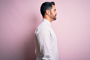 Sakallı, rahat gömlekli genç bir adam pembe arka planda dikiliyor, yan yana bakıyor, rahat poz veriyor, kendine güvenen bir gülümsemesi var..