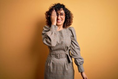 Güzel bir Arap iş kadını elbise ve gözlük takıyor sarı arka planda duruyor bir gözünü eliyle kapatıyor, yüzünde kendine güvenen bir gülümseme ve sürpriz bir duygu var..