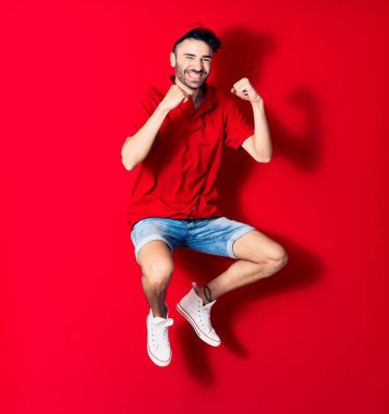 Şapkalı genç teslimatçı mutlu gülümsüyor. Yüzünde tebessümle zıplıyor, kırmızı arka planda yumruklarıyla kazanan jestler yapıyor.