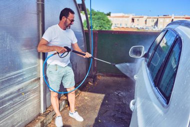 Orta yaşlı yakışıklı, günlük kıyafetler giyen ve mutlu gülümseyen güneş gözlüklü bir adam. Yüzünde gülümsemeyle ayakta durup su tabancası kullanarak araba yıkamak..