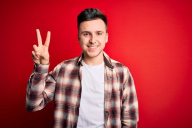 Kırmızı izole edilmiş arka plan üzerine gündelik modern tişört giyen yakışıklı beyaz adam iki numarayı gösterirken kendine güvenen ve mutlu bir şekilde gülümsüyor..