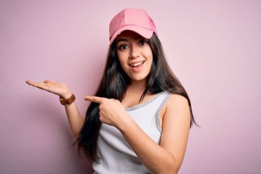 Pembe arka planda spor şapkalı genç esmer kadın hayrete düşmüş ve kameraya gülümsüyor..