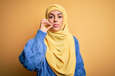 Genç, güzel, esmer, Müslüman kadın izole edilmiş sarı arka plan ağzı üzerine Arap başörtüsü takıyor ve dudakları parmaklarıyla kapanıyor. Gizli ve sessiz, tabu konuşması