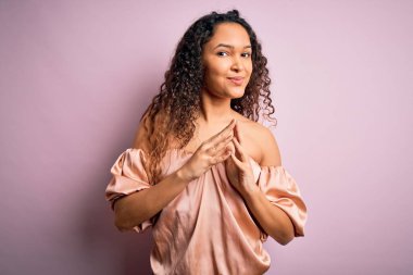 Kıvırcık saçlı, pembe tişörtlü genç ve güzel bir kadın pembe arka planda el ele tutuşup gülümsüyor ve rahatlıyor. Başarı ve iyimser