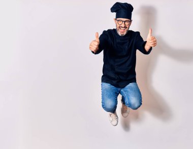 Orta yaşlı, yakışıklı, gözlüklü ve aşçı üniformalı mutlu bir adam. Yüzünde tebessümle zıpla, tamam işareti yap, başparmağınla izole beyaz arkaplan üzerinde.