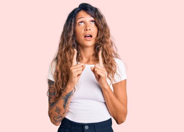 Gündelik beyaz tişört giyen genç İspanyol kadın şaşırmış ve şaşırmış bir şekilde yukarı bakıp parmaklarını göstermiş ve kollarını kaldırmıştı.. 