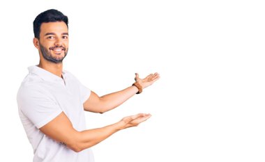 Genç, yakışıklı, günlük kıyafetler giyen, gülümseyerek içeri girmeye davet eden, açık elle gülümseyen bir İspanyol. 