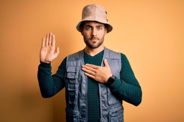 Tatilde sakallı yakışıklı turist sarı arka planda kaşif şapkası takıyor göğsü ve avucu açık yemin ediyor sadakat yemini ediyor.