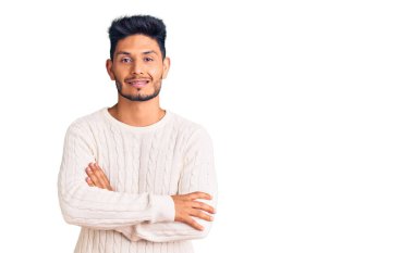 Yakışıklı Latin Amerikalı genç adam sıradan bir kazak giyip kollarını kavuşturup kameraya bakarken gülümsüyor. pozitif kişi. 