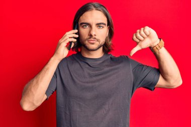 Genç yakışıklı bir adam akıllı telefonda konuşurken sinirli bir yüz ifadesi, olumsuz işaret baş parmakları aşağıda, reddedilme konsepti. 