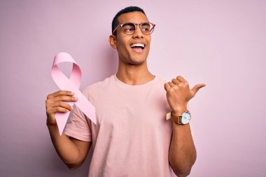 Genç ve yakışıklı Afro-Amerikalı adam pembe kanser kurdelesini soyutlanmış arka planda tutuyor ve gülümseyen bir gülümsemeyle baş parmağıyla işaret ediyor.