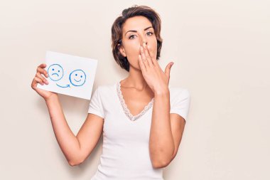 Genç, güzel bir kadın, elinde üzgün ve mutlu bir duygu kağıdıyla ağzını eliyle kapatıyor, şok geçiriyor ve hata yapmaktan korkuyor. şaşırmış ifade 