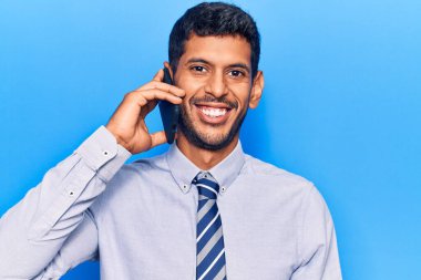 Genç Latin adam akıllı telefondan konuşuyor. Olumlu ve mutlu görünüyor. Kendine güvenen bir gülümsemeyle dişlerini gösteriyor. 