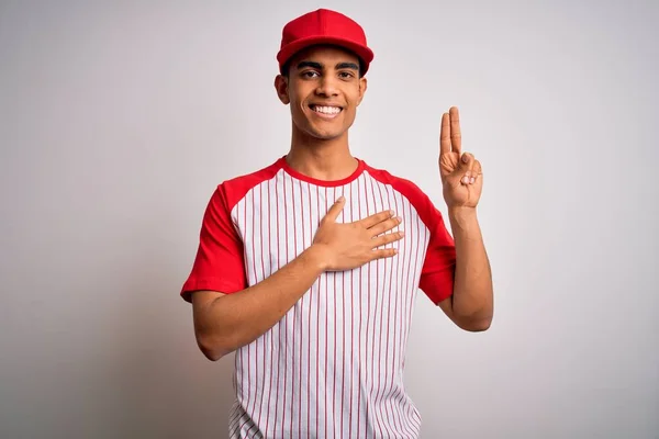 若いハンサムなアフリカ系アメリカ人のスポーツマンは 縞模様の野球Tシャツとキャップを着て胸と指で手で身に着けている笑顔 忠誠の誓いをする — ストック写真