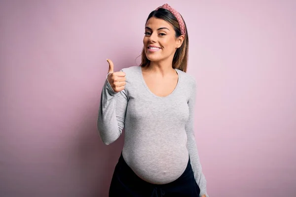 孤立したピンクの背景に赤ちゃんを期待若い美しいブルネットの女性は手で幸せな親指アップジェスチャーを行う上で妊娠 成功を示すカメラを見て表現を承認する — ストック写真