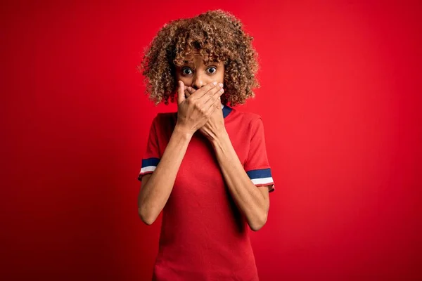 年轻美丽的非洲女人穿着休闲的T恤 站在红色的背景上 双手捂住嘴 为自己的错误而震惊 秘密概念 — 图库照片