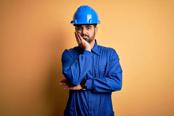 Mechanic man with beard wearing blue uniform and safety helmet over yellow background thinking looking tired and bored with depression problems with crossed arms.