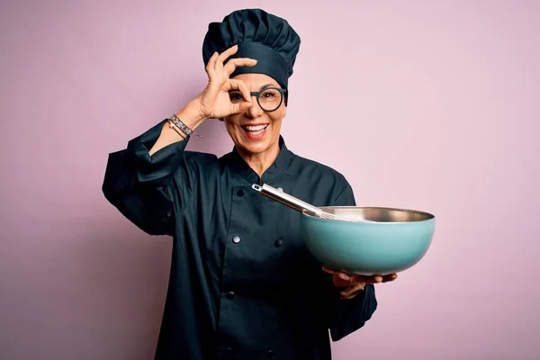Middle age brunette chef woman wearing cooker uniform and hat using whisk and bowl with happy face smiling doing ok sign with hand on eye looking through fingers