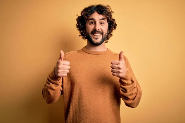黄色の背景成功サインの上に立ってカジュアルなセーターを着てひげを持つ若いハンサムな男は 手で積極的なジェスチャーを行うと 親指を笑顔と幸せ 陽気な表情と勝者のジェスチャー — ストック写真