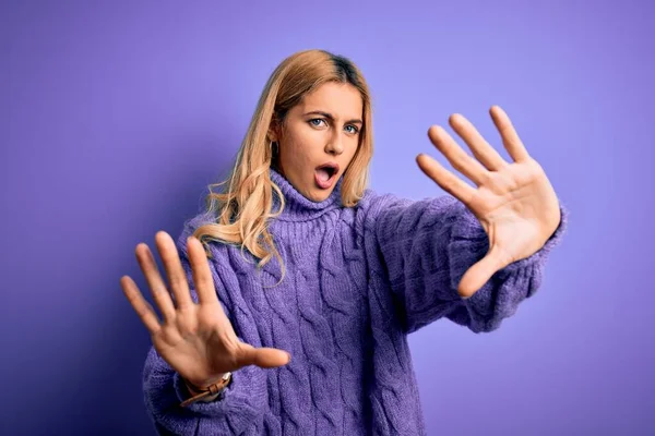 Young beautiful blonde woman wearing casual turtleneck sweater over purple background doing stop gesture with hands palms, angry and frustration expression