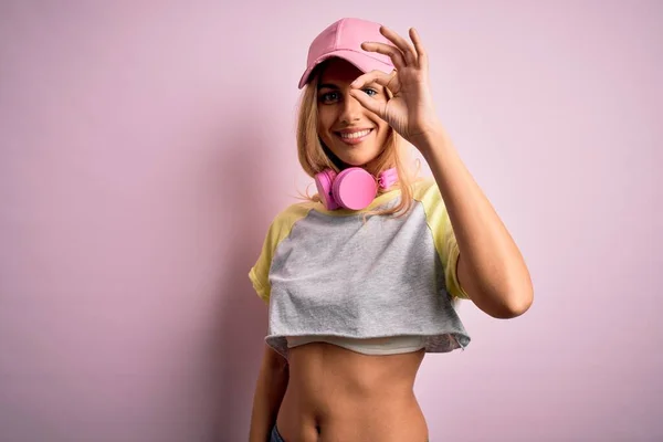 Young beautiful blonde sportswoman doing sport using headphones over pink background doing ok gesture with hand smiling, eye looking through fingers with happy face.