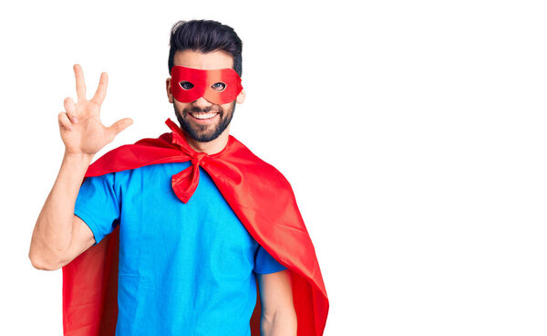 Young handsome man with beard wearing super hero costume showing and pointing up with fingers number three while smiling confident and happy. 