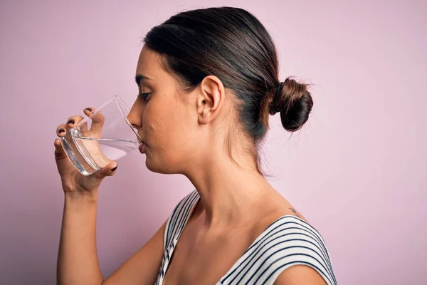 若いです美しいですブルネット女性飲むガラスの健康的な水へリフレッシュスタンディング上の隔離されたピンクの背景 — ストック写真
