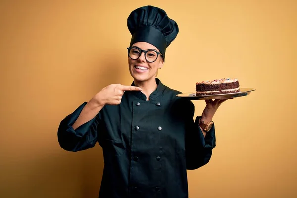 Young beautiful brunette baker woman wearing cooker uniform and hat holding cake very happy pointing with hand and finger
