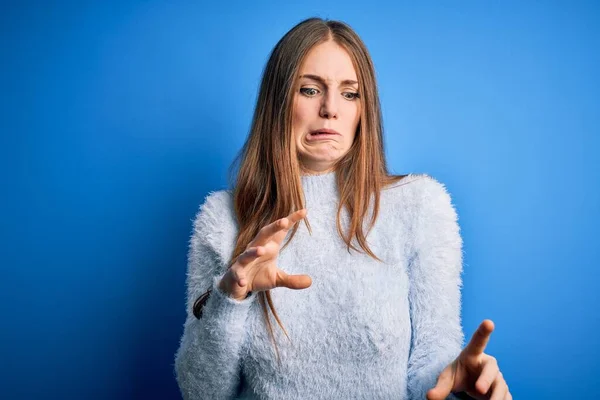 Young beautiful redhead woman wearing casual sweater over isolated blue background disgusted expression, displeased and fearful doing disgust face because aversion reaction.