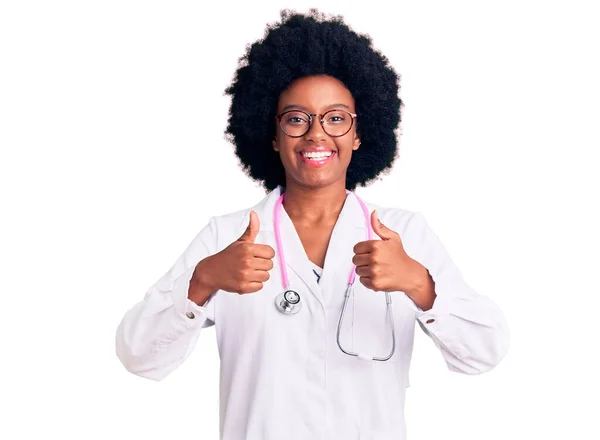 医師のコートと聴診器の成功のサインを身に着けている若いアフリカ系アメリカ人女性は 手で積極的なジェスチャーを行います 親指を笑顔と幸せ 陽気な表情と勝者のジェスチャー — ストック写真