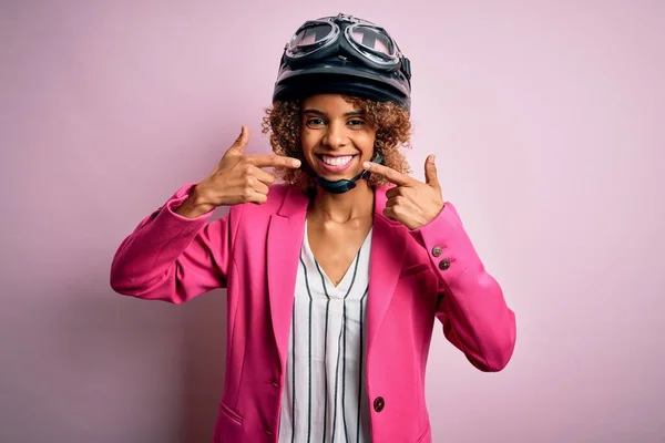 アフリカ系アメリカ人のオートバイの女性は ピンクの背景に元ヘルメットを身に着けている巻き毛が陽気で 指の歯と口を指して笑っています 歯科衛生の概念 — ストック写真