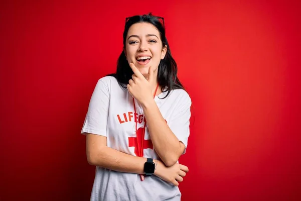 Young lifeguard woman wearing secury guard equipent over red background looking confident at the camera smiling with crossed arms and hand raised on chin. Thinking positive.