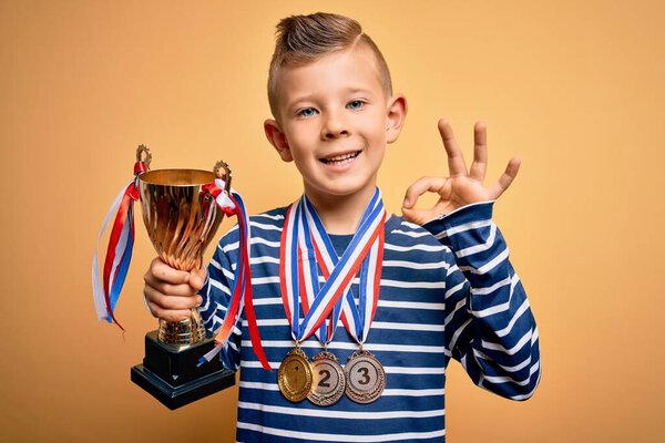 Маленький кавказский паренек с медалями победителя и трофеем за победу на жёлтом фоне делает знак ОК с пальцами, отличный символ
