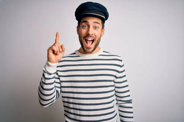 Young handsome sailor man with beard wearing navy striped uniform and captain hat pointing finger up with successful idea. Exited and happy. Number one.