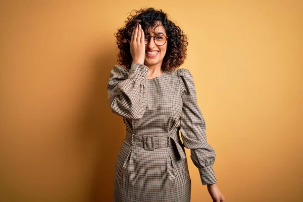 Güzel bir Arap iş kadını elbise ve gözlük takıyor sarı arka planda duruyor bir gözünü eliyle kapatıyor, yüzünde kendine güvenen bir gülümseme ve sürpriz bir duygu var..