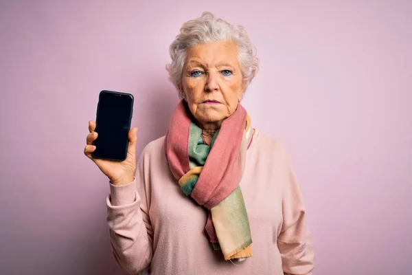 ピンクを背景にスマートフォンの画面を持つシニア美しい白髪の女性が真剣に考えてスマート顔に自信を持って表現 — ストック写真