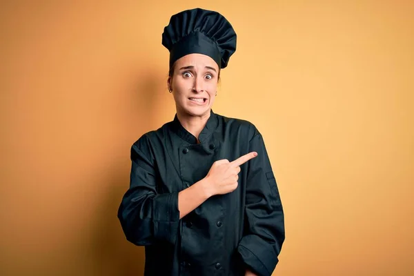 Young beautiful chef woman wearing cooker uniform and hat standing over yellow background Pointing aside worried and nervous with forefinger, concerned and surprised expression