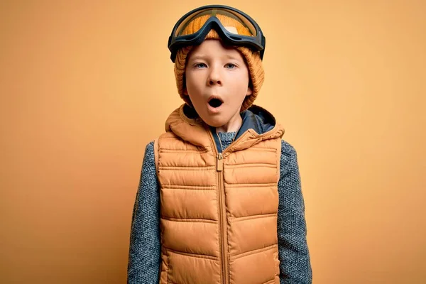 Young little caucasian kid wearing snow glasses and winter coat over yellow background afraid and shocked with surprise expression, fear and excited face.