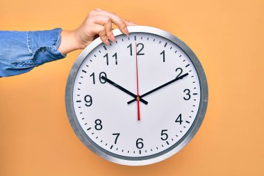 Sarı arka planda büyük saati tutan güzel bir kadın.